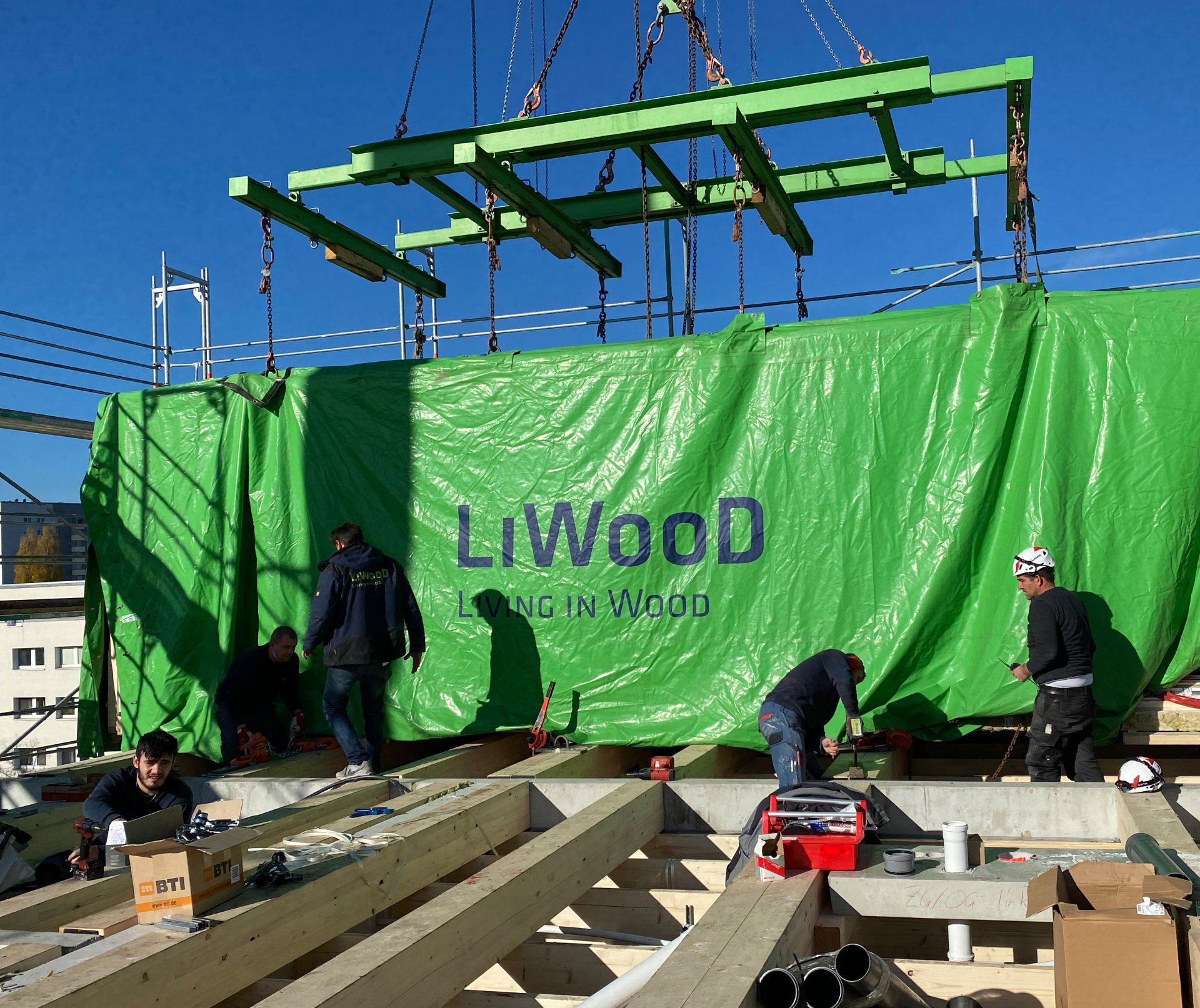 Several men working on a construction site in nice weather. In the background, there is a green tarpaulin with the inscription ‘Living in Wood’.